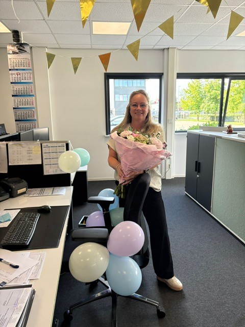 Medewerker met een boeket bloemen bij een versierde werkplek met ballonnen.